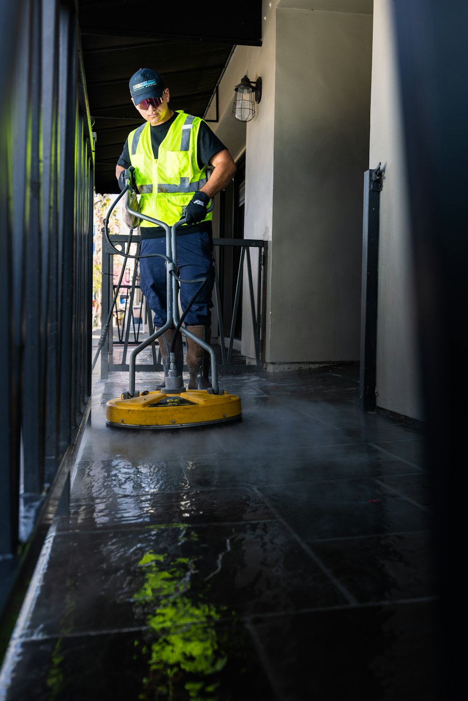 Un homme en gilet haute visibilité nettoie à haute pression un sol carrelé extérieur.